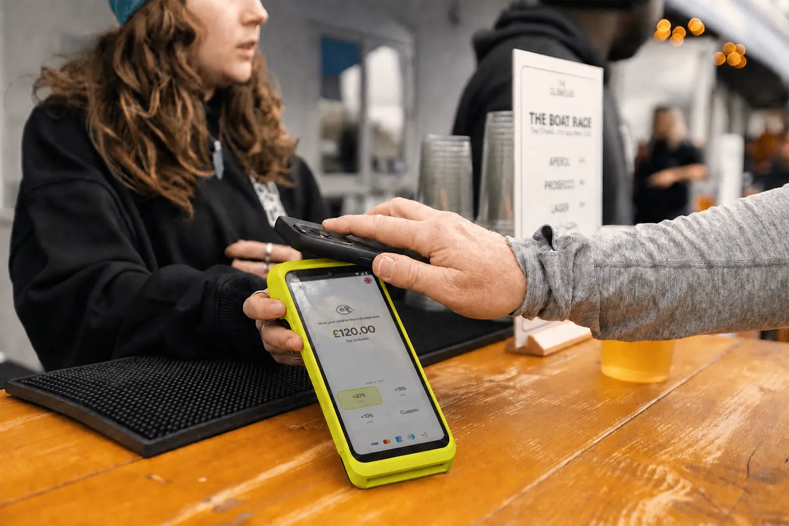 A barista handing a Teya Pro terminal across a wooden bar counter while a customer taps a phone on it.